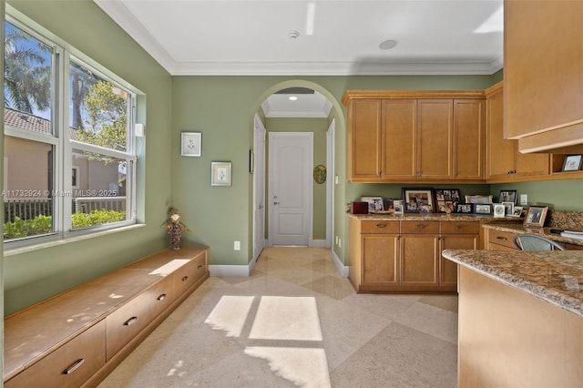 kitchen with baseboards, arched walkways, light stone counters, brown cabinets, and ornamental molding