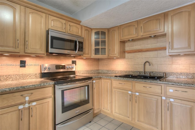 kitchen with light tile patterned floors, light brown cabinets, a sink, appliances with stainless steel finishes, and glass insert cabinets