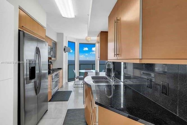 kitchen featuring light tile patterned floors, stainless steel appliances, a sink, brown cabinetry, and dark stone countertops