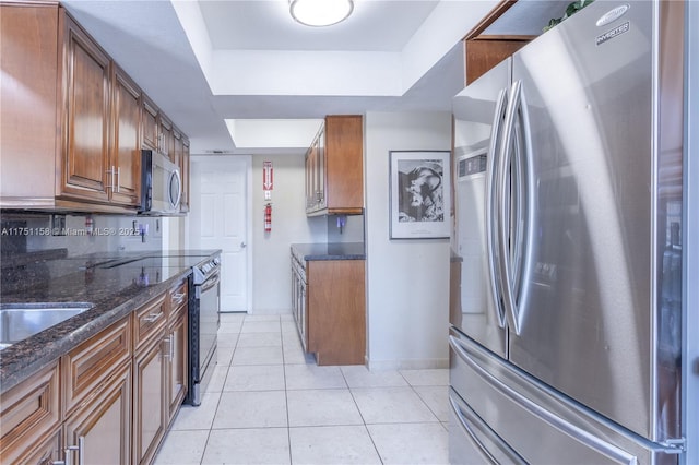 kitchen with a tray ceiling, appliances with stainless steel finishes, brown cabinetry, light tile patterned flooring, and dark stone countertops