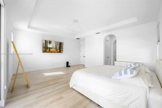 bedroom with arched walkways, a tray ceiling, baseboards, and light wood-style floors