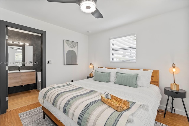 bedroom featuring light wood finished floors, ensuite bathroom, a ceiling fan, a sink, and baseboards