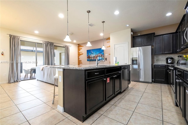 kitchen featuring an island with sink, light stone counters, hanging light fixtures, stainless steel appliances, and dark cabinetry