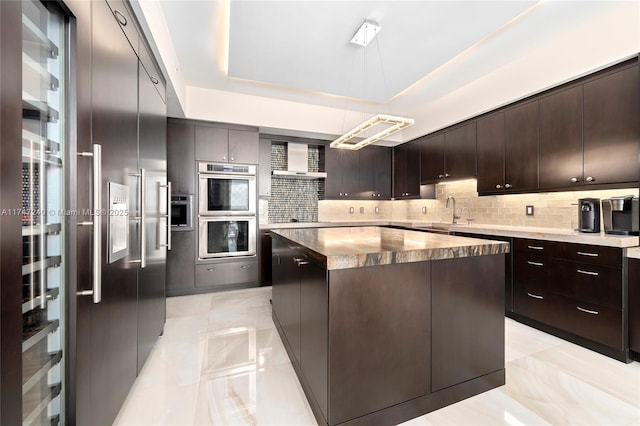 kitchen with dark brown cabinetry, stainless steel appliances, a kitchen island, backsplash, and a tray ceiling