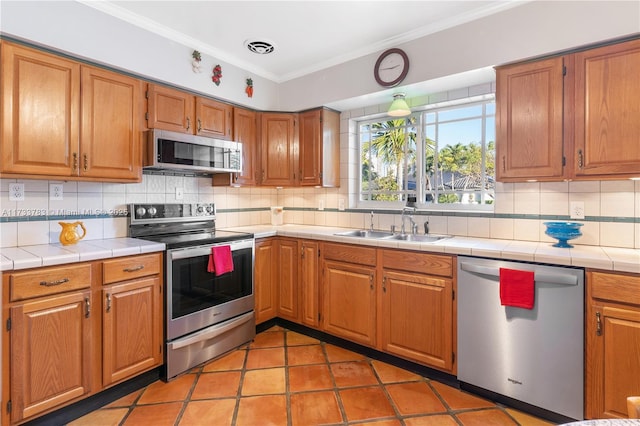 kitchen with tile counters, appliances with stainless steel finishes, brown cabinets, ornamental molding, and a sink