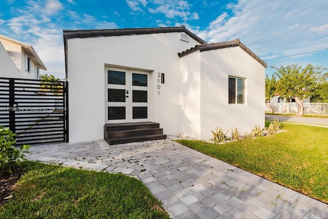 entrance to property featuring fence, a tiled roof, french doors, a lawn, and stucco siding