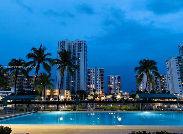 pool at twilight with a view of city lights, fence, and a community pool