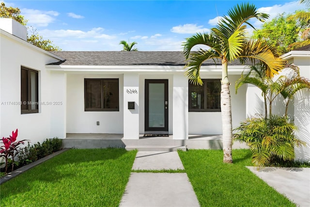 view of exterior entry featuring a shingled roof, a porch, a lawn, and stucco siding