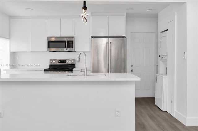 kitchen featuring stainless steel appliances, hanging light fixtures, white cabinets, a sink, and stacked washing maching and dryer