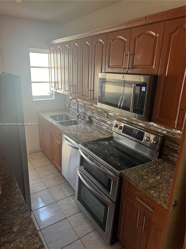 kitchen with stainless steel appliances, a sink, decorative backsplash, brown cabinets, and dark stone countertops