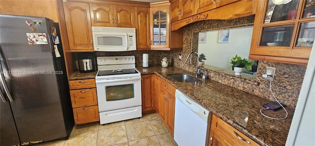 kitchen featuring brown cabinetry, glass insert cabinets, a sink, dark stone countertops, and white appliances