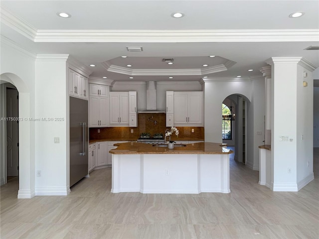 kitchen featuring arched walkways, a tray ceiling, stainless steel built in fridge, an island with sink, and wall chimney exhaust hood