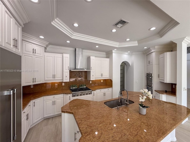 kitchen with an island with sink, wall chimney exhaust hood, white cabinetry, and a raised ceiling