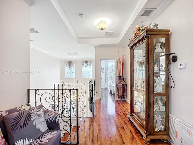 corridor featuring visible vents, a tray ceiling, wood finished floors, and an upstairs landing