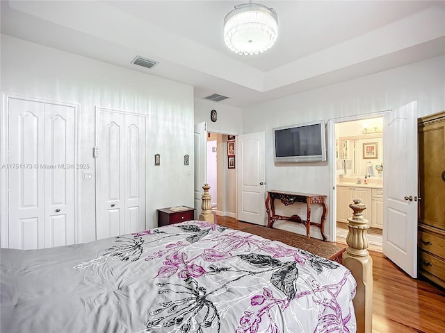 bedroom with two closets, ensuite bath, visible vents, and wood finished floors