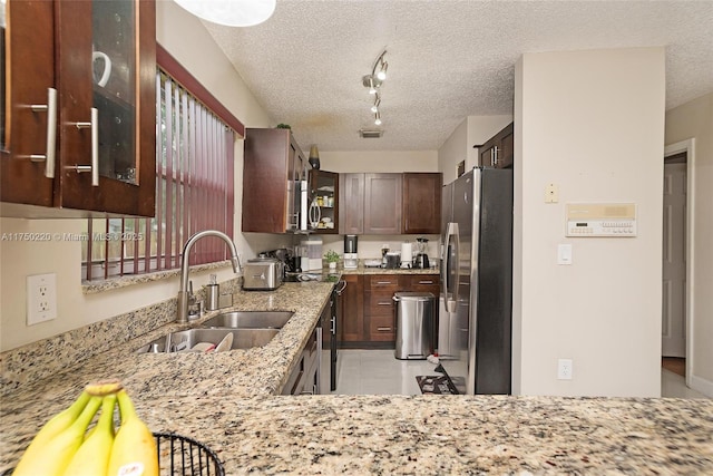 kitchen featuring a textured ceiling, stainless steel appliances, a sink, light stone countertops, and glass insert cabinets