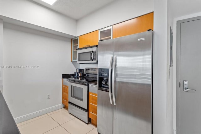 kitchen featuring light tile patterned floors, baseboards, appliances with stainless steel finishes, dark countertops, and glass insert cabinets