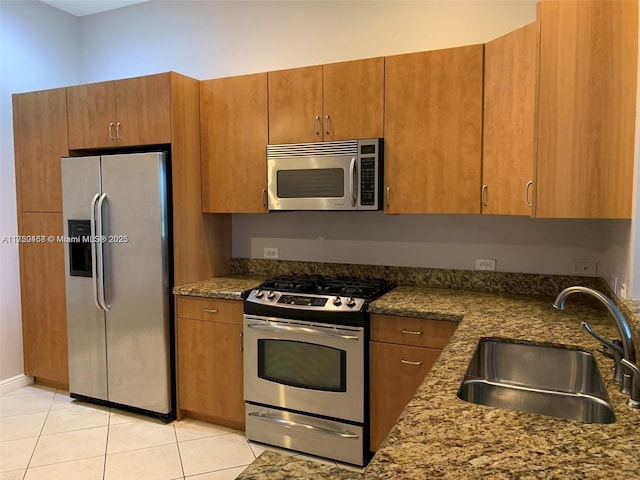 kitchen featuring appliances with stainless steel finishes, brown cabinets, dark stone countertops, a sink, and light tile patterned flooring