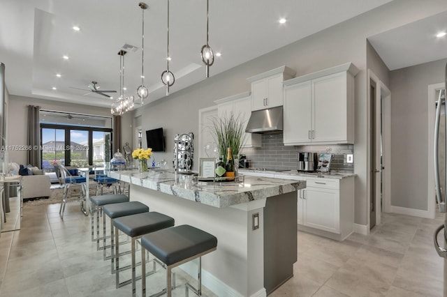 kitchen featuring light stone counters, tasteful backsplash, a raised ceiling, open floor plan, and under cabinet range hood