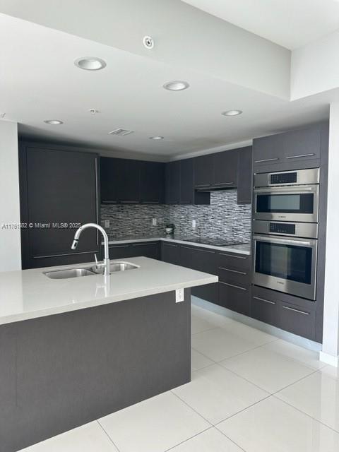 kitchen featuring decorative backsplash, dark cabinets, light countertops, stainless steel double oven, and a sink