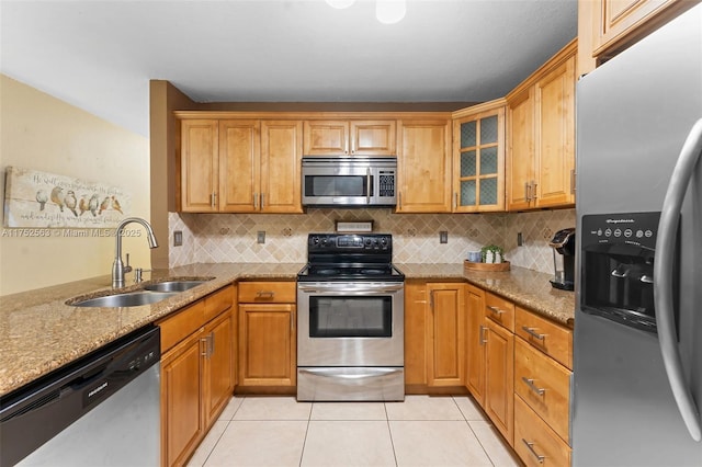 kitchen with stainless steel appliances, brown cabinetry, glass insert cabinets, a sink, and light stone countertops