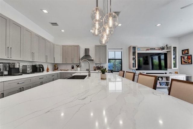 kitchen featuring gray cabinetry, a kitchen breakfast bar, wall chimney range hood, light stone countertops, and pendant lighting