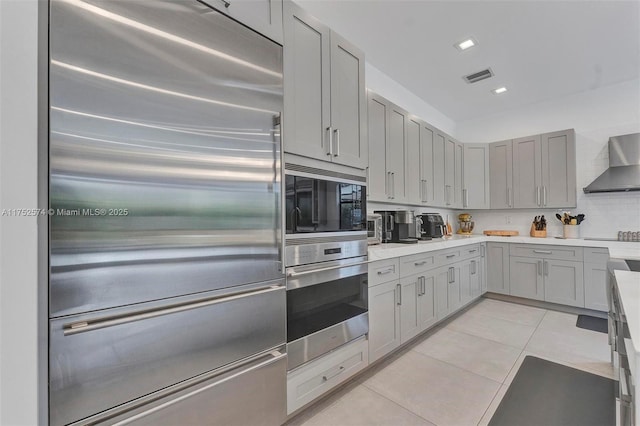 kitchen with light tile patterned floors, visible vents, built in appliances, light countertops, and wall chimney range hood