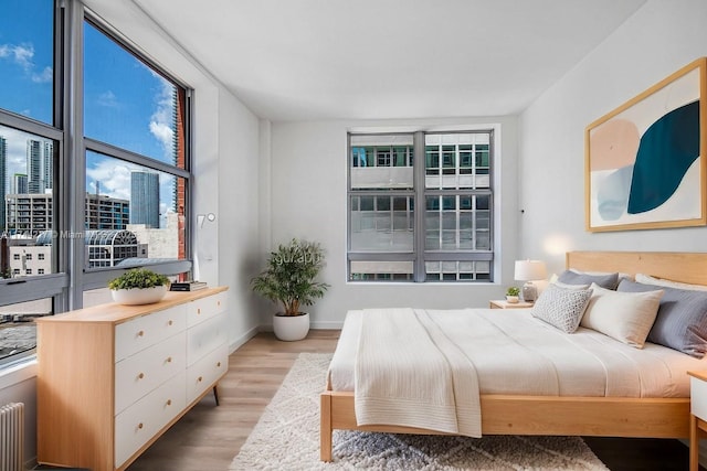 bedroom featuring a view of city, radiator heating unit, light wood-type flooring, and baseboards