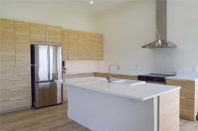 kitchen featuring wall chimney exhaust hood, appliances with stainless steel finishes, light brown cabinetry, light wood-style floors, and a sink