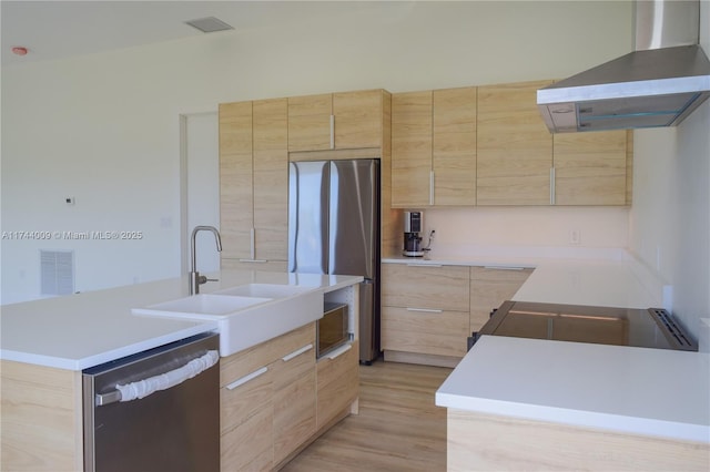 kitchen featuring light brown cabinetry, appliances with stainless steel finishes, a sink, modern cabinets, and wall chimney exhaust hood