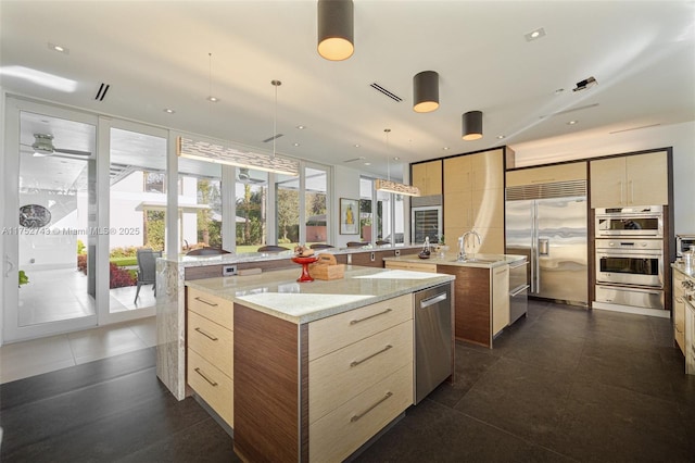 kitchen featuring light stone counters, a kitchen island with sink, stainless steel appliances, light brown cabinetry, and a warming drawer