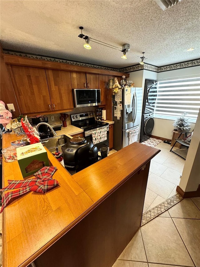 kitchen featuring a textured ceiling, light tile patterned flooring, stainless steel appliances, stacked washing maching and dryer, and brown cabinetry