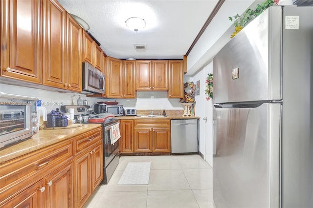 kitchen with brown cabinets, stainless steel appliances, light tile patterned flooring, a sink, and light stone countertops