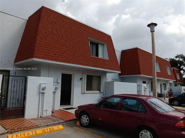 view of front facade with stucco siding, uncovered parking, mansard roof, and roof with shingles