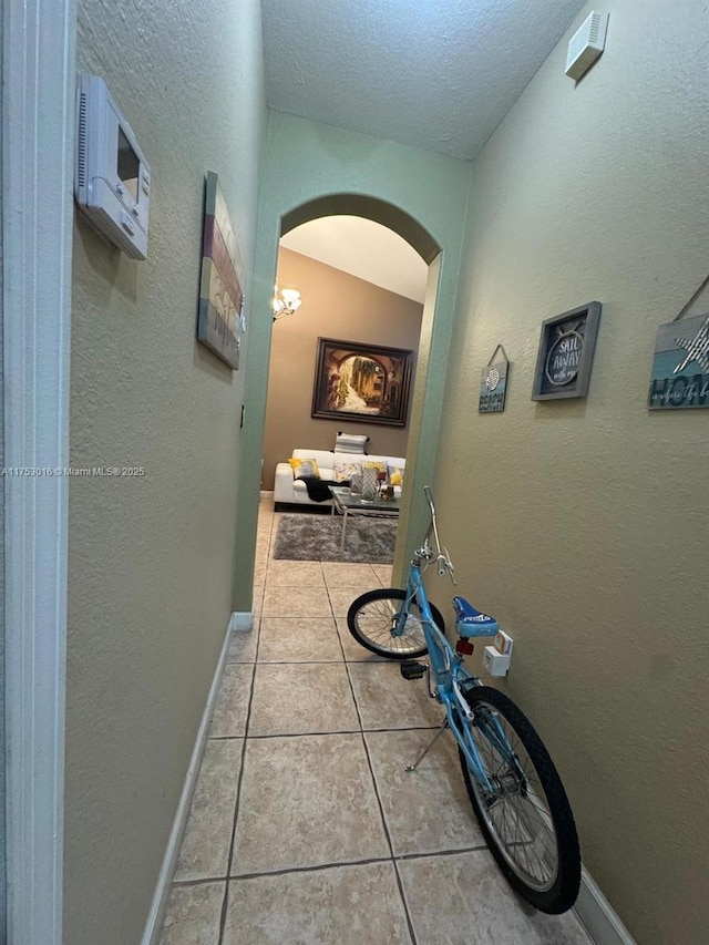 hallway featuring arched walkways, a textured wall, light tile patterned floors, a textured ceiling, and baseboards