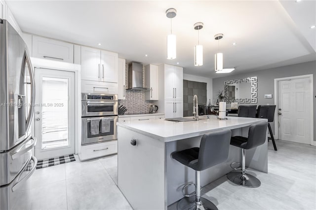 kitchen featuring decorative light fixtures, stainless steel appliances, white cabinets, an island with sink, and wall chimney exhaust hood