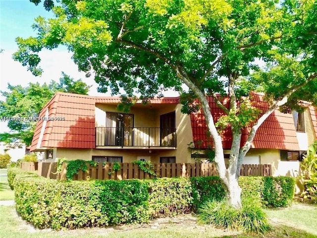 rear view of house featuring mansard roof, a balcony, a tile roof, fence, and stucco siding