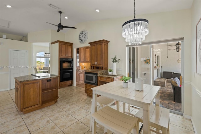 kitchen with light tile patterned flooring, dobule oven black, ceiling fan with notable chandelier, brown cabinetry, and stainless steel microwave