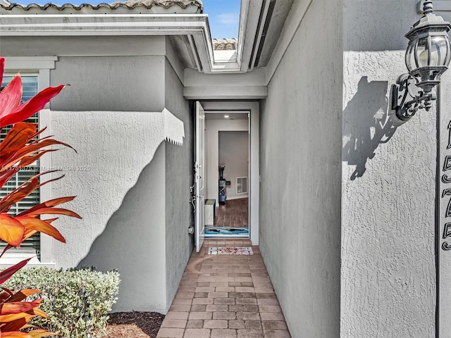 entrance to property with a tiled roof, visible vents, and stucco siding