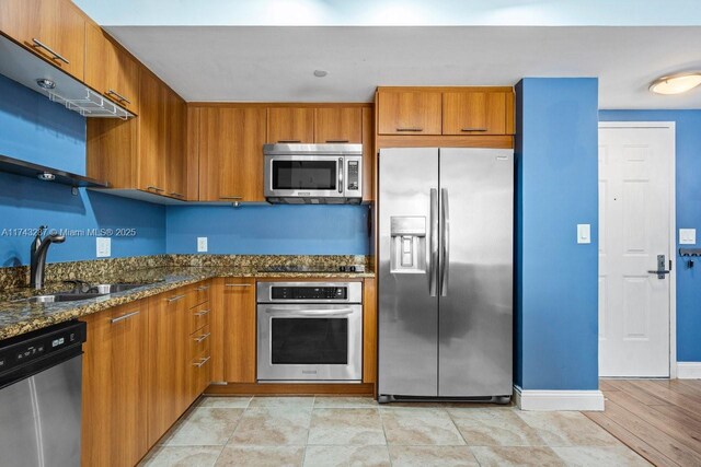 kitchen featuring a sink, baseboards, appliances with stainless steel finishes, brown cabinets, and dark stone counters