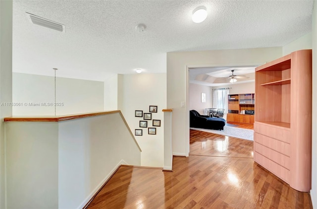 hallway with light wood-style floors, visible vents, a textured ceiling, and an upstairs landing