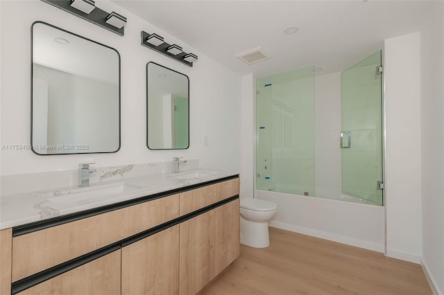 bathroom with double vanity, bath / shower combo with glass door, toilet, wood finished floors, and a sink