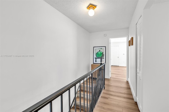 hall with light wood finished floors, baseboards, a textured ceiling, and an upstairs landing