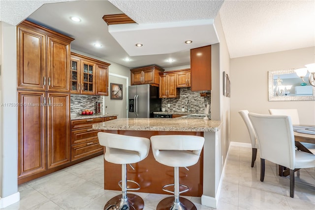 kitchen featuring glass insert cabinets, brown cabinets, a peninsula, a kitchen bar, and stainless steel refrigerator with ice dispenser