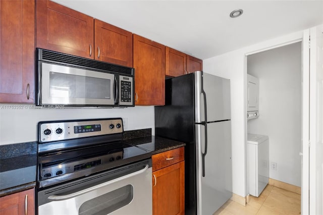 kitchen featuring light tile patterned floors, stacked washer / dryer, appliances with stainless steel finishes, brown cabinets, and dark stone countertops