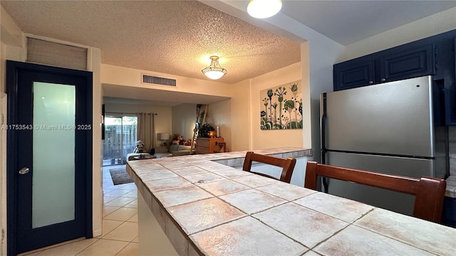 kitchen with light tile patterned flooring, visible vents, blue cabinetry, tile counters, and freestanding refrigerator