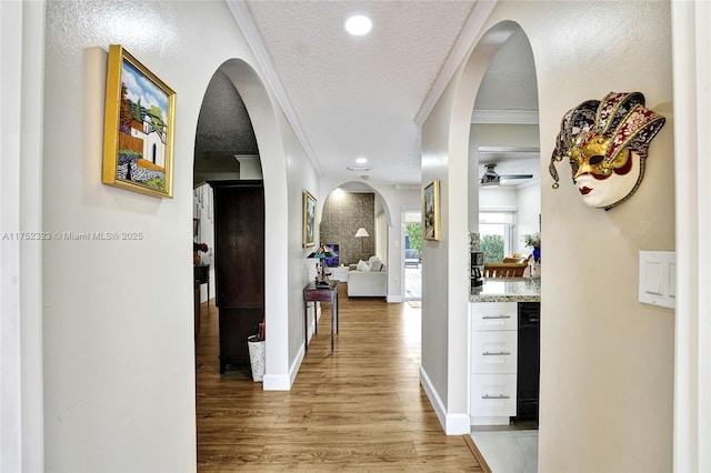 hall featuring arched walkways, a textured ceiling, baseboards, light wood finished floors, and crown molding