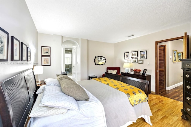 bedroom featuring arched walkways, visible vents, a textured ceiling, light wood-type flooring, and baseboards