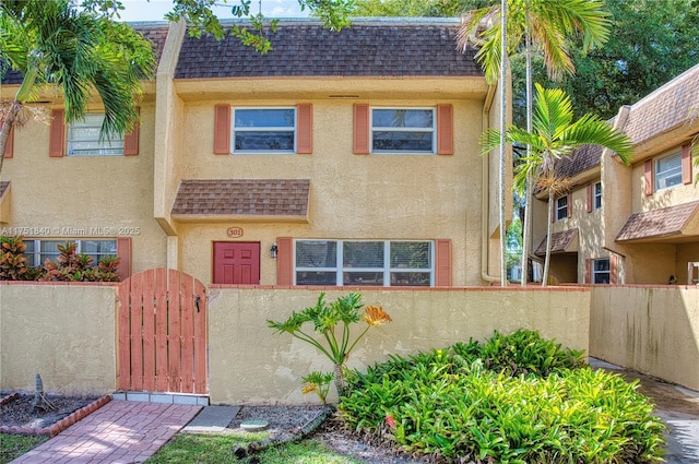 multi unit property featuring a shingled roof, a gate, mansard roof, and stucco siding