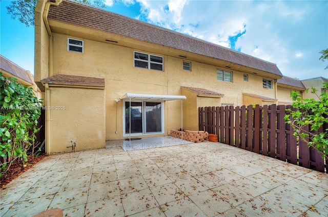 rear view of property with mansard roof, fence, roof with shingles, stucco siding, and a patio area